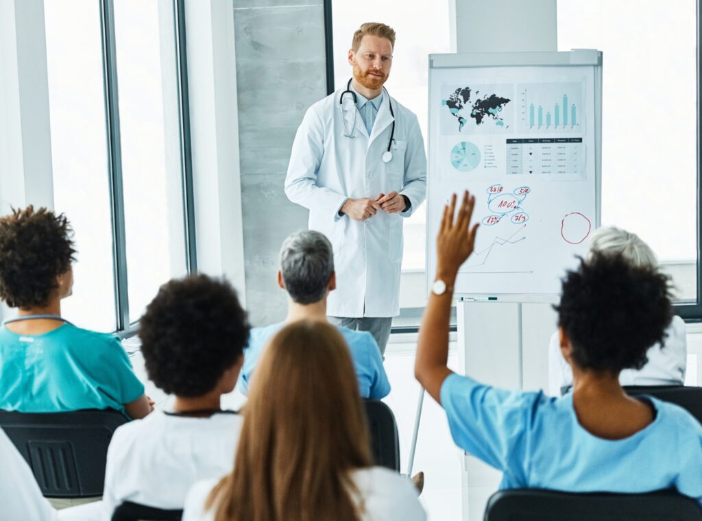 Image shows a doctor wearing a lab coat and stethoscope around his neck standing next to a display easel and in front of a group of attendees. The attendees are sitting in chairs and have their back to the camera listening intently to the doctor’s presentation. One person has their hand up to represent asking a question.